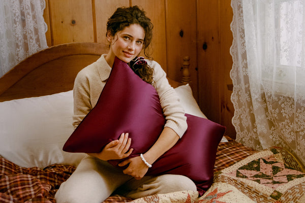 A woman sits on a bed in a rustic bedroom setting, holding a crimson/burgundy silk pillowcase. She is wearing a cream-colored cardigan and pants.