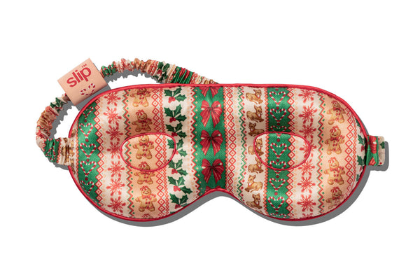 A festive Christmas-themed contour sleep mask by Slip, featuring a patterned fabric with gingerbread men, snowflakes, holly, and bows in green, red, and beige, with a matching patterned strap.