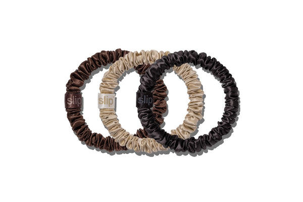 Three pure silk hair scrunchies in dark brown, beige, and black, overlapping on a clean white background. Each scrunchie features a small fabric brand tag with the word 'slip'.