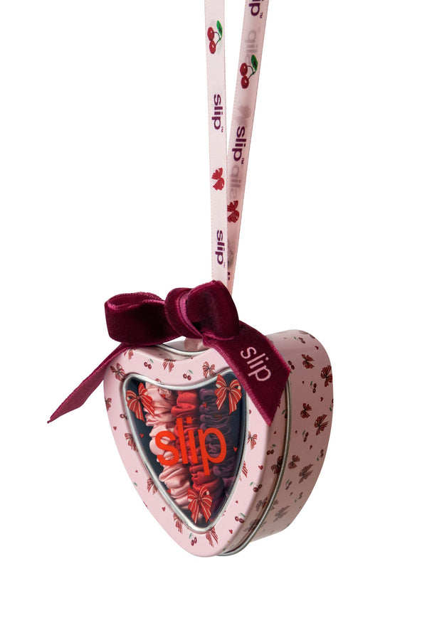 A heart-shaped Slip tin with a pink cherry pattern, a dark red velvet bow, and a hanging ribbon printed with the Slip brand name.