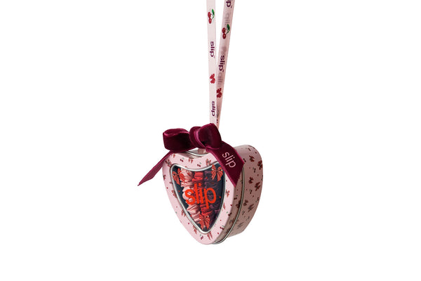 A heart-shaped Slip tin with a pink cherry pattern, a dark red velvet bow, and a hanging ribbon printed with the Slip brand name.