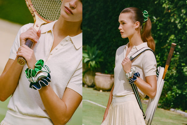 Woman holding a vintage tennis racket on a tennis court with greenery in the background and slip silk scrunchies on her wrist
