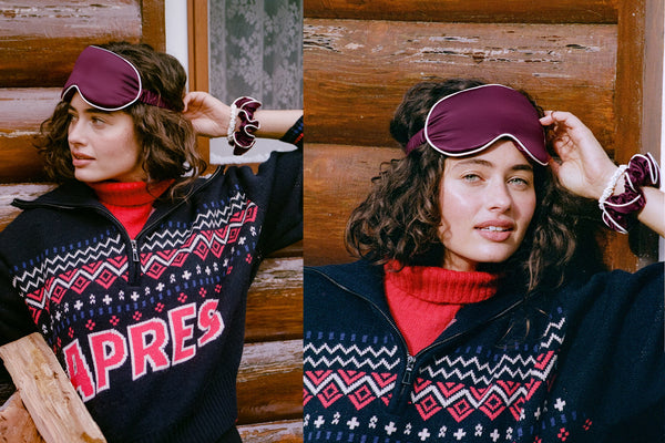 A split-screen lifestyle image showing a woman wearing a burgundy silk contour sleep mask, a red sweater, and a navy blue patterned zip-up. She is posed against a rustic wooden log background.