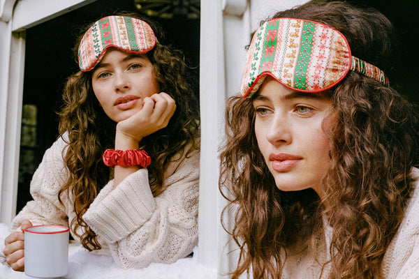 A woman wearing a festive holiday slip silk sleep mask with a red, green, and cream striped pattern, styled in a cozy lifestyle setting with a white knit sweater and a red silk scrunchie.