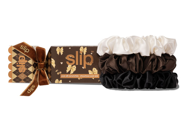 Three slip large silk scrunchies in white, chocolate, and black are displayed next to their branded packaging cracker box on a clean white background. The box is brown with gold patterns and a brown velvet bow.