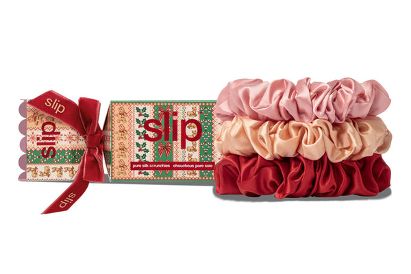 Three slip silk hair scrunchies in pink, peach, and red are displayed next to their Christmas-themed packaging box with a red ribbon, on a white background.
