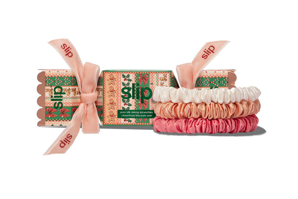 Three pure silk skinny scrunchies in white, peach and pink, displayed in front of their patterned gift box, tied with a pink ribbon printed with the brand name 'slip'.