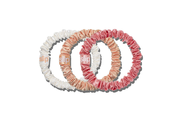 Three slip silk skinny hair scrunchies in white, peach, and pink, arranged in a circular overlapping pattern on a solid white background. The brand name 'slip' is visible on the tag of each scrunchie.