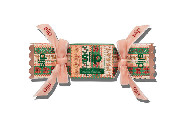 A scrunchie cracker package tied with a pink ribbon, shown on a plain white background. The wrapper has a holiday pattern in green, red, and beige, with the brand name 'slip' visible on the ribbon and wrapper.