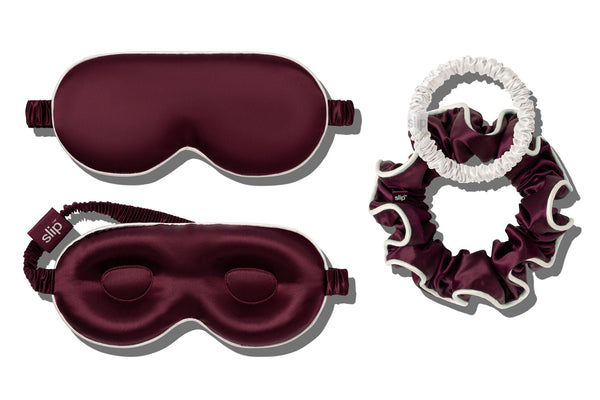 A sleep and haircare set featuring a crimson contour sleep mask with white piping, and matching large silk scrunchie, and a white skinny scrunchie, arranged on a white background.