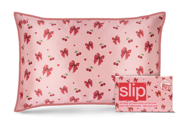 A pink Slip brand silk pillow with a red bow and cherry pattern and  lace trim, shown next to its matching pink packaging box on a white background.