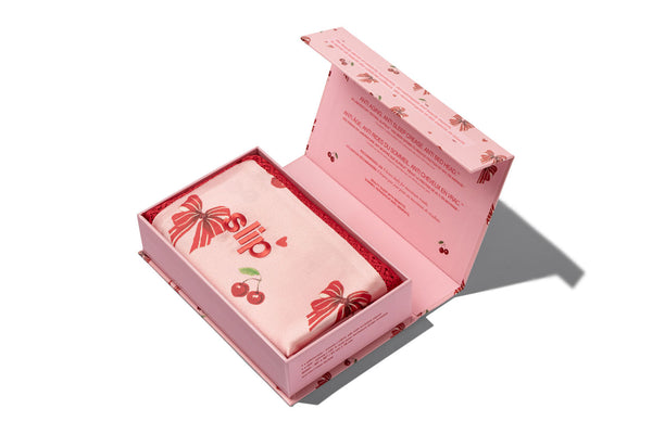 A slip silk pillowcase box with its lid open, displaying a folded pillowcase printed with red bows and cherries, set against a clean white background.
