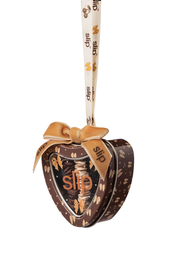 A heart-shaped metal gift tin with a hanging ribbon, featuring a bow design and the brand name 'slip' printed on the lid and ribbon, set against a plain white background.