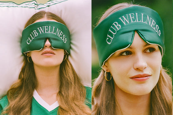 Two women wearing green 'Club Wellness' silk sleep masks with a blurred background.